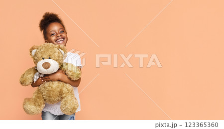 A little black girl with dark hair is smiling brightly as she holds a large, brown teddy bear in front of a peach-colored background. The girl is wearing a white t-shirt and blue jeans, copy space 123365360