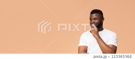 A young black man with a beard is smiling and looking off to the side with his hand on his chin. He is wearing a white t-shirt and is standing in front of a light brown background, copy space A young black man with a beard is smiling and looking off to the side with his hand on his chin. He is wearing a white t-shirt and is standing in front of a light brown background, copy space 123365368