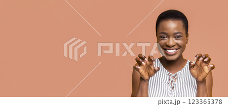 African American young woman with short, dark hair is smiling brightly. She is standing against a peach-colored backdrop. Both of her hands are held up in a playful, clawing gesture 123365378