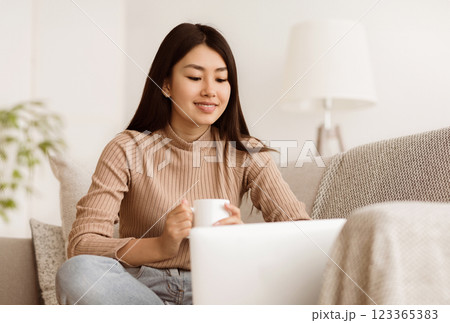 A woman is sitting on a couch in her home, using a laptop and drinking coffee. She is dressed casually and looks relaxed. 123365383