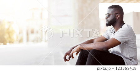 A young man with a beard and sunglasses sits by a window in a contemplative pose. 123365419