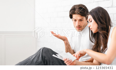 A young couple sits on a couch in their living room, looking at paperwork and talking. They are likely discussing their finances, perhaps bills or budgeting. The woman points to the documents 123365431