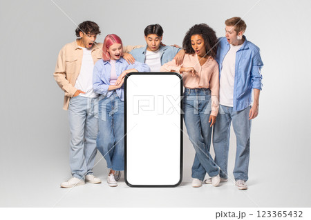 A group of five young adults, all dressed in casual attire, stand in front of a large smartphone with a blank white screen. They are all looking at the phone with expressions of interest, excitement 123365432