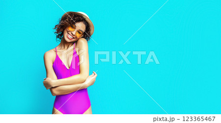 This image features African American smiling woman with curly brown hair wearing a pink swimsuit and a white hat. She is posing in front of a bright blue background, copy space 123365467