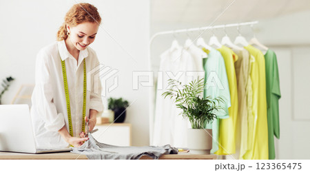 A young woman in a white shirt cuts fabric at a table in a home studio. She is wearing a measuring tape and is smiling. A young woman in a white shirt cuts fabric at a table in a home studio. She is wearing a measuring tape and is smiling. 123365475