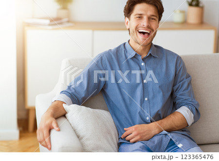 A man in a blue button-down shirt sits on a white couch and laughs. He is relaxed and looks happy. A man in a blue button-down shirt sits on a white couch and laughs. He is relaxed and looks happy. 123365476
