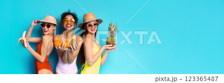 Three young women wearing swimsuits and summer hats are holding various fruits, including a banana, oranges, and a pineapple. The women are smiling and posing against blue, copy space 123365487
