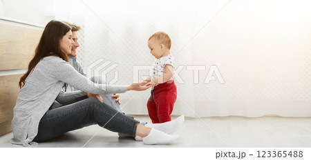 A young family of three are spending time together at home. The parents are sitting on the floor, smiling at their baby who is taking their first steps, copy space 123365488