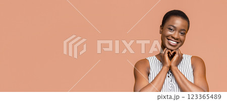 African American young woman with short hair is smiling and looking directly at the camera. She is making a heart shape with her hands in front of her chest 123365489