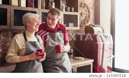 A happy senior couple stands together in their kitchen, both holding red mugs of coffee. They are smiling and laughing as they share a moment together, empty space 123365518