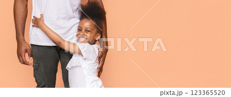 A little African American girl with curly hair is hugging her father from behind. She is smiling and looking directly at the camera. Her father is standing to the right of the frame, copy space A little African American girl with curly hair is hugging her father from behind. She is smiling and looking directly at the camera. Her father is standing to the right of the frame, copy space 123365520