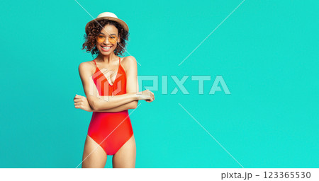 A young African American woman is wearing a red swimsuit and straw hat, with a bright, toothy smile on her face. She stands against a turquoise background, with her arms crossed, copy space A young African American woman is wearing a red swimsuit and straw hat, with a bright, toothy smile on her face. She stands against a turquoise background, with her arms crossed, copy space 123365530