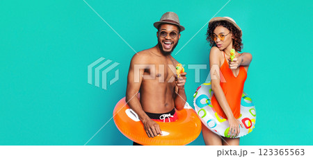 A happy couple is posing against a bright teal background. The man is wearing a brown fedora and sunglasses and holding a yellow water gun. He is holding an orange inflatable ring, copy space A happy couple is posing against a bright teal background. The man is wearing a brown fedora and sunglasses and holding a yellow water gun. He is holding an orange inflatable ring, copy space 123365563