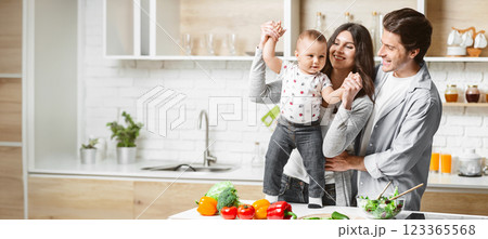 A happy family is playing in their modern kitchen while preparing food. The mother is holding up their young child, who is smiling and reaching out. Father is standing next to them, looking lovingly 123365568