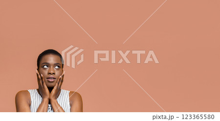 Black young woman with short hair looking upwards in surprise or wonder. She is wearing a light-colored striped top and has her hands clasped near her face. The background is a peach-colored wall. 123365580