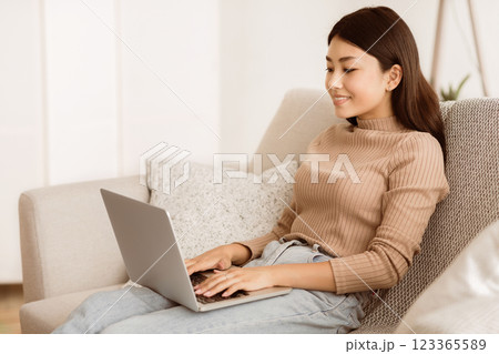 A young woman sits on a couch in a living room, working on her laptop computer. She is smiling and appears to be focused on her work. 123365589