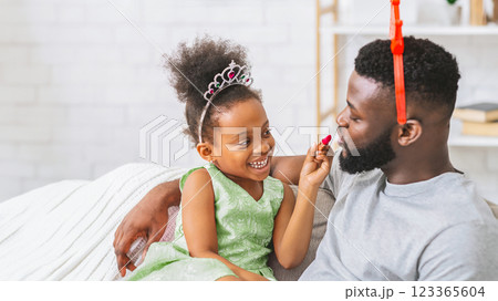 African American young girl with a princess crown is putting lipstick on her fathers face. She is smiling and having fun while her father sits patiently and looks at her. 123365604