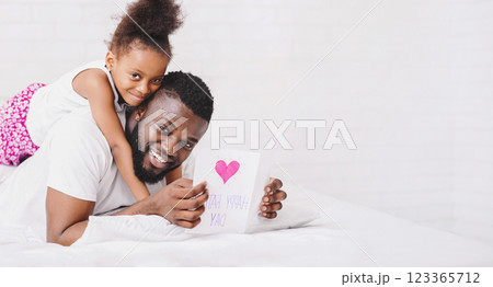 Black father is lying in bed holding a card that has a pink heart and handwritten words on it, smiling at the camera. His daughter is sitting on his back, her arms wrapped around his neck 123365712