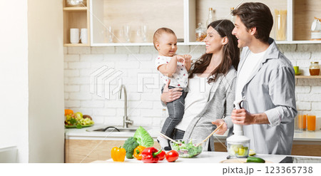 A young family is preparing a healthy meal together in a modern kitchen. The mother is holding their baby while the father prepares food with a handheld blender, copy space 123365738