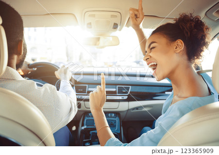 African American woman is enthusiastically singing along to the music in the passenger seat of a car. She is raising her hand in the air and has a big smile on her face 123365909