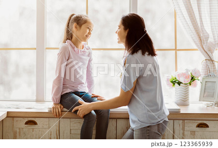 A young girl sits on a kitchen counter while a woman stands next to her, both looking at each other and talking. The window behind them is open, and there is a vase of pink flowers on the counter 123365939