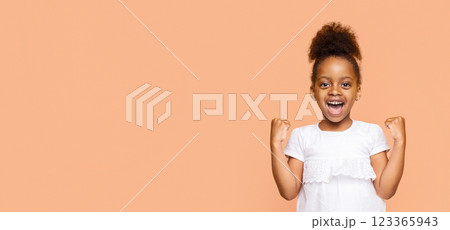 A little Black girl with a big smile and her fists raised in the air celebrates a victory or achievement. She is wearing a white shirt and her dark hair is styled in a high ponytail, copy space 123365943