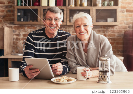 A happy senior couple smiles for the camera while sitting at a kitchen table, enjoying coffee and using a tablet. The man is wearing a striped shirt, and the woman is wearing a grey sweater. 123365966