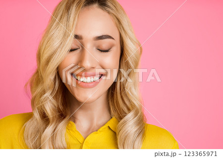 A young woman with long blonde hair smiles while looking down. She is wearing a yellow shirt and has her eyes closed. The background is a bright pink. A young woman with long blonde hair smiles while looking down. She is wearing a yellow shirt and has her eyes closed. The background is a bright pink. 123365971