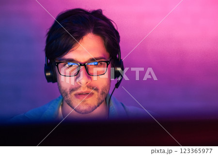 A young man with dark hair and a beard is wearing glasses and headphones, focused on a computer screen. The room is lit with a purple and pink gradient light, creating a vibrant and modern aesthetic 123365977