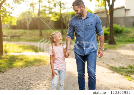 A father and his young daughter walk hand-in-hand on a gravel path through a park. The sun shines brightly, casting long shadows on the ground. They are both smiling, enjoying the beautiful day 123365983
