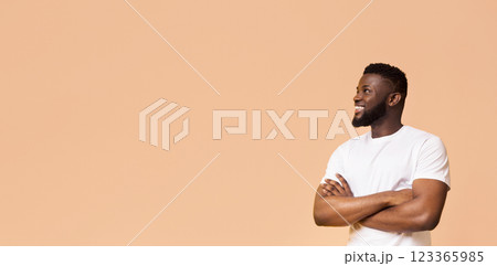 A smiling black man stands against a peach background with his arms crossed. He is wearing a white t-shirt and looking off to the side. The man is confident and relaxed, copy space A smiling black man stands against a peach background with his arms crossed. He is wearing a white t-shirt and looking off to the side. The man is confident and relaxed, copy space 123365985