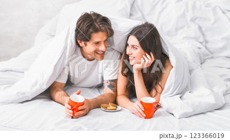 A couple, a man and a woman, are lying in bed, covered by white bedding. They are both smiling and looking at each other, holding mugs of coffee and a plate of cookies. A couple, a man and a woman, are lying in bed, covered by white bedding. They are both smiling and looking at each other, holding mugs of coffee and a plate of cookies. 123366019