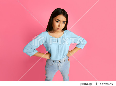 Asian young woman standing with her hands on her hips, looking disappointed. She is wearing a light blue blouse and light blue jeans. The background is a solid pink color. 123366025