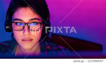 A close-up shot of a young woman with dark hair wearing glasses and headphones, looking intently at a computer screen. The background is a vibrant mix of purple and blue lighting, copy space 123366047
