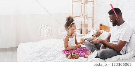 African American father and his little daughter are sitting on a bed, both wearing playful headbands. They are smiling and looking at each other, engaged in a game with stuffed toys. African American father and his little daughter are sitting on a bed, both wearing playful headbands. They are smiling and looking at each other, engaged in a game with stuffed toys. 123366063