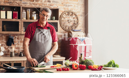 A man, wearing glasses and an apron, is smiling as he cuts vegetables on a cutting board in a modern kitchen. He is surrounded by a variety of colorful vegetables 123366065