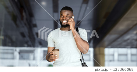 A man smiles while talking on the phone and holding a cup of coffee. He is walking outdoors and wearing a white t-shirt. A man smiles while talking on the phone and holding a cup of coffee. He is walking outdoors and wearing a white t-shirt. 123366069