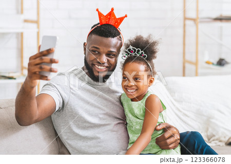 A happy black father and daughter, both wearing crowns, sit on the couch together and take a selfie with a smartphone. The father is smiling and holding the phone out in front of him. A happy black father and daughter, both wearing crowns, sit on the couch together and take a selfie with a smartphone. The father is smiling and holding the phone out in front of him. 123366078