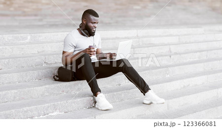 A man sits on concrete stairs in an urban setting, working on a laptop while holding a coffee cup. 123366081