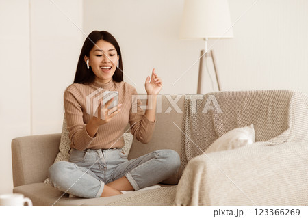 Asian Girl Listening Music in Airpods and Singing Song on Sofa, Free Space Asian Girl Listening Music in Airpods and Singing Song on Sofa, Free Space 123366269