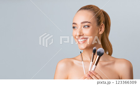 A young woman with long blonde hair pulled back into a ponytail is holding three makeup brushes in front of a light grey background. She is smiling and looking at copy space A young woman with long blonde hair pulled back into a ponytail is holding three makeup brushes in front of a light grey background. She is smiling and looking at copy space 123366309