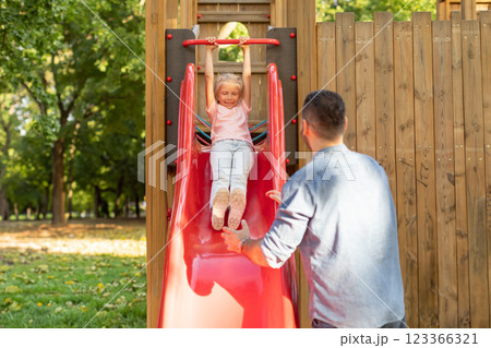 A young girl in a pink shirt and blue jeans slides down a red slide on a playground. Her father stands below the slide watching her. He has his hand out as if he is ready to catch her 123366321