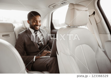 African American man in a business suit is seated in the back seat of a car, working on a laptop. He is smiling and appears to be focused on his work African American man in a business suit is seated in the back seat of a car, working on a laptop. He is smiling and appears to be focused on his work 123366329
