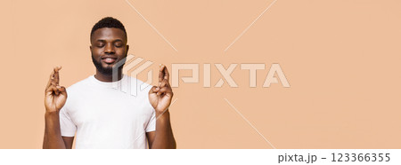 African American man with crossed fingers, praying with his eyes closed against a peach-colored background. The man is wearing a white t-shirt, and his expression is serene, copy space African American man with crossed fingers, praying with his eyes closed against a peach-colored background. The man is wearing a white t-shirt, and his expression is serene, copy space 123366355