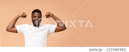 A happy, young, Black man with a short beard is flexing his muscles while standing in front of a beige background. He is wearing a white t-shirt and has a big smile on his face, copy space, panorama 123366362