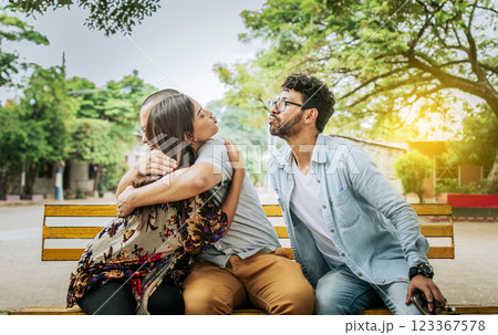 Unfaithful woman sitting hugging her boyfriend and secretly kissing another man. Unfaithful girlfriend hugging her boyfriend in a park secretly kissing another man, Couple infidelity concept 123367578