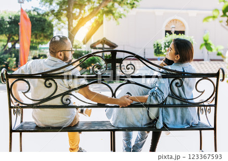 Cheating girlfriend with her boyfriend holding another man hand in a park. Unfaithful woman sitting holding hands with another man while boyfriend hugs her. love triangle concept 123367593