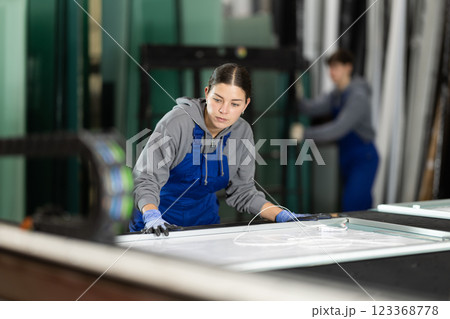 Girl glazer employee in blue overall works in glass processing shop Girl glazer employee in blue overall works in glass processing shop 123368778