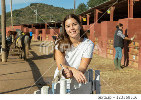 Portrait of cheerful European young woman wearing white T-shirt and posing on yard of horse club 123369316
