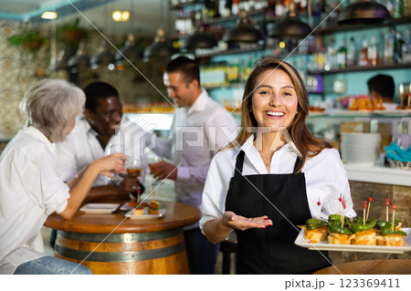 Happy Latin female waiter in uniform pointing hand on the plate with pinchos behind a table with diverse friends and bar counter 123369411